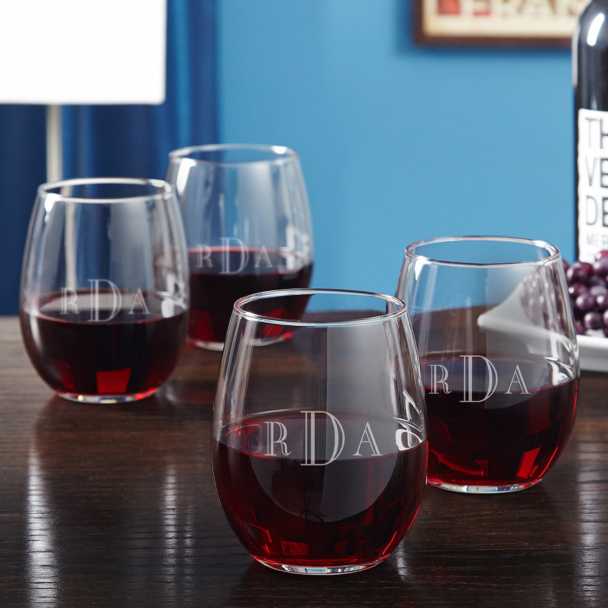 Four stemless wine glasses with red wine and monogram letters on a wooden table, bottle and grapes in background