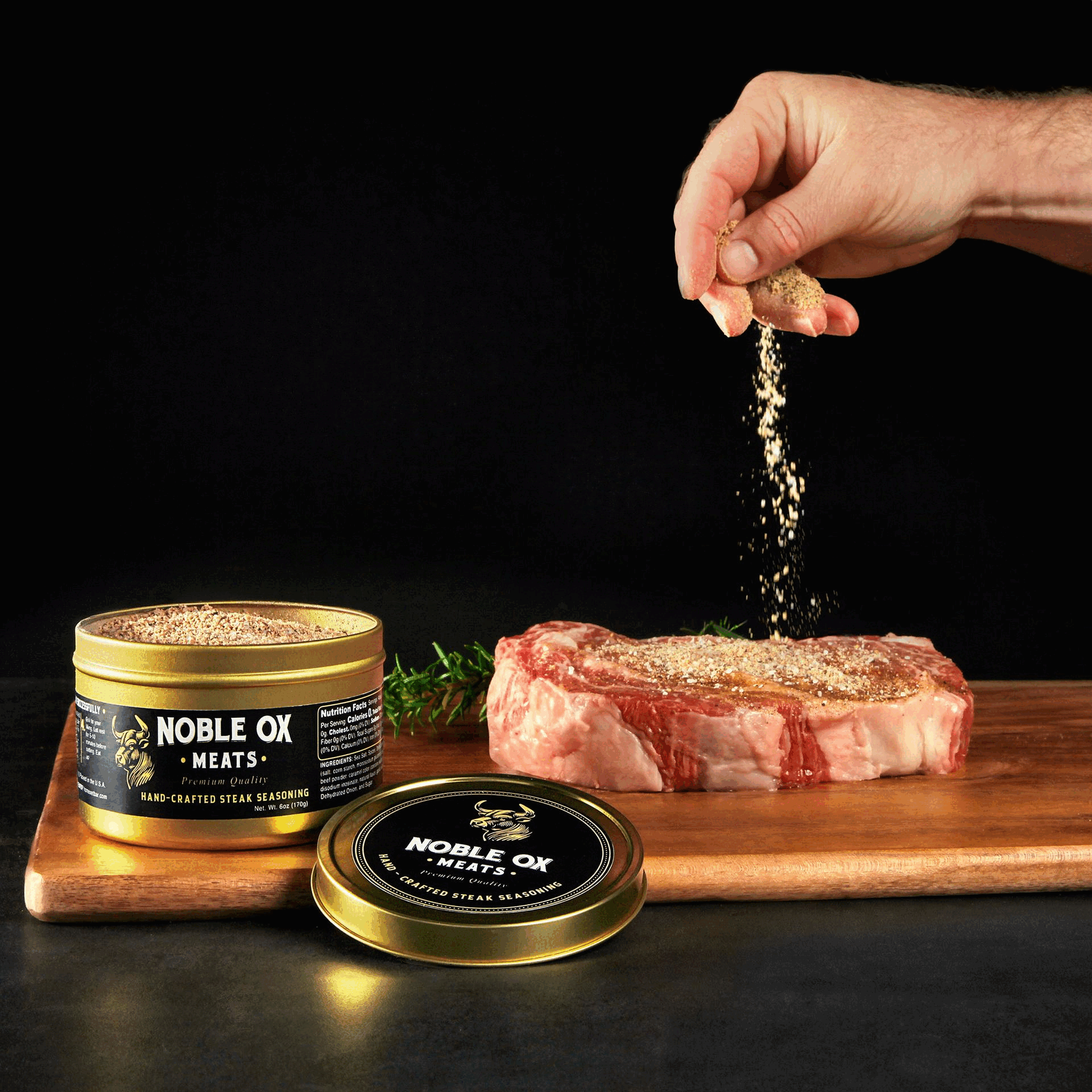 Hand sprinkling seasoning onto a raw steak on a cutting board beside an open tin labeled “Noble Ox Meats”.