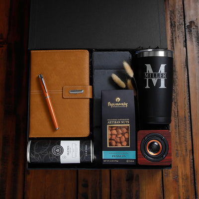 Gift box with leather notebook, orange pen, snack pack, beverage can, personalized black tumbler, and small speaker.