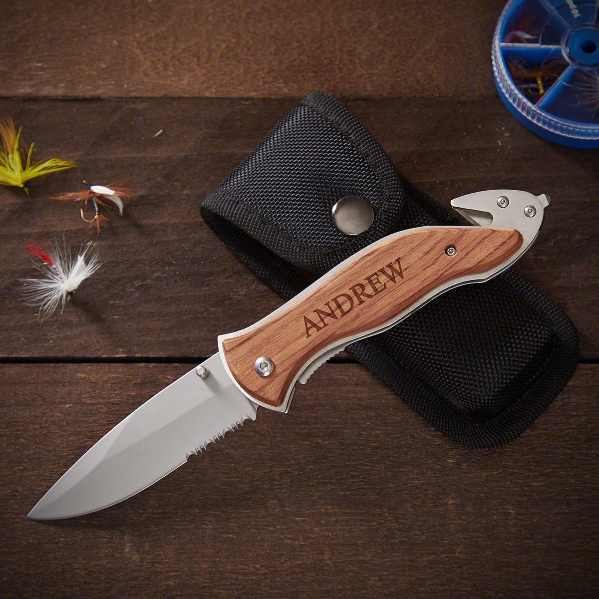Folding knife with wooden handle engraved “ANDREW” resting on black pouch, with fishing flies on wooden table.