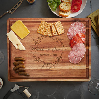 Engraved charcuterie board reading “Matt & Jenny” and “October 16, 2027” with meats, cheese, crackers, and pickles.