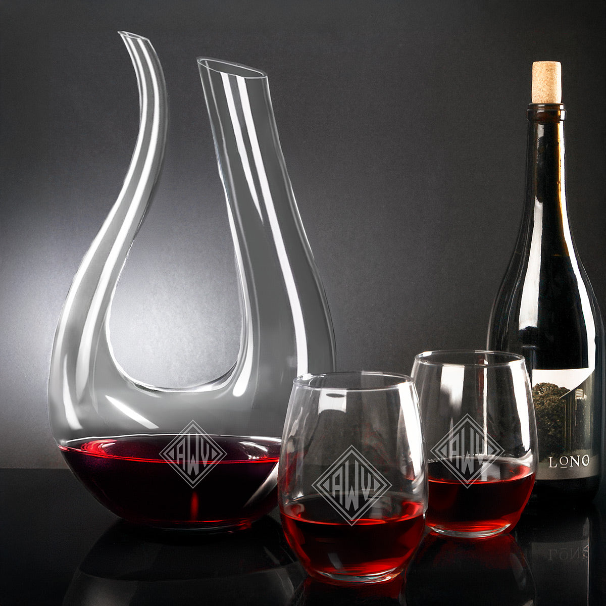 Glass wine decanter with red wine, two stemless glasses, and a corked wine bottle on a dark background.