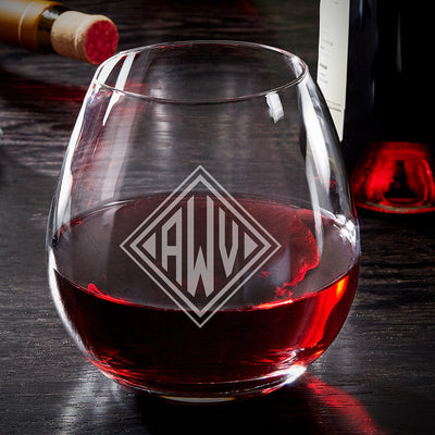 Stemless wine glass half-filled with red wine, etched with “CAW” monogram; wine bottle and cork in background.
