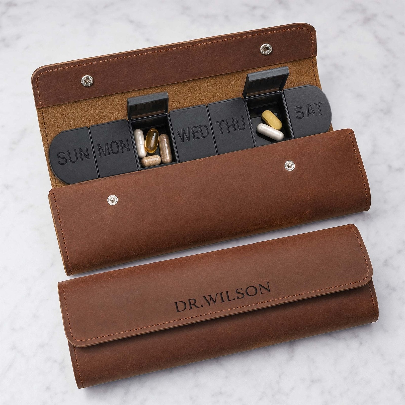 Open leather weekly pill case with labeled compartments and capsules on a marble surface