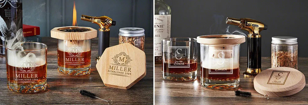 Two whiskey glasses on a table with a torch, smoking wood chip glass, jar of wood chips, and bottle in background.