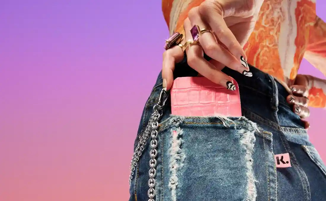 Hand with rings places a pink textured wallet into the back pocket of distressed jeans with a chain.