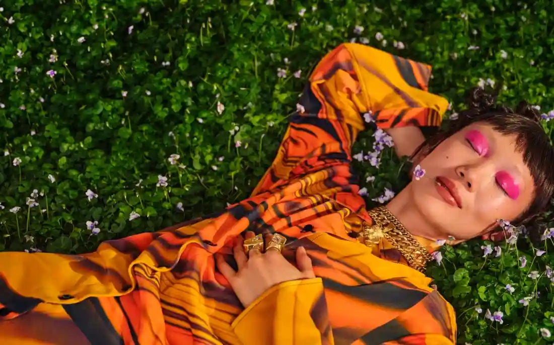 Person lying on green grass with small flowers, wearing orange patterned outfit and pink eyeshadow.