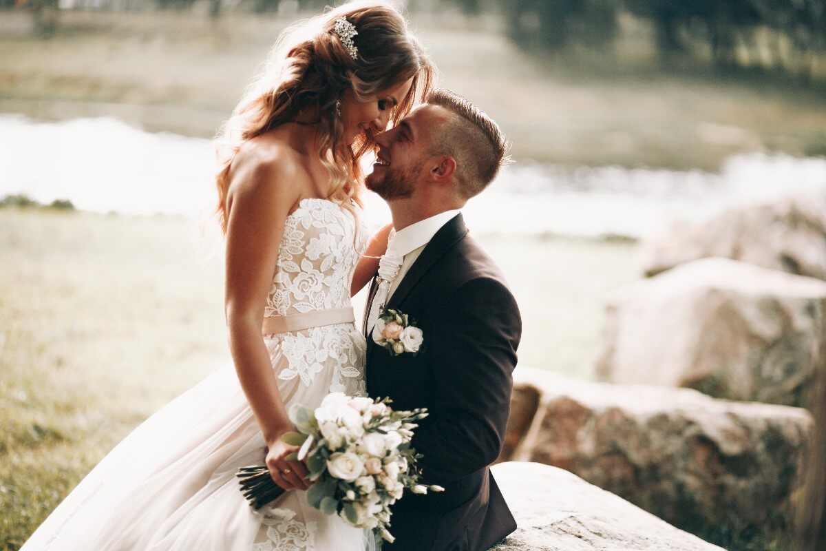 Bride in lace gown holding bouquet leans toward groom in black suit outdoors near rocks and water.