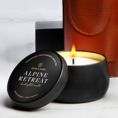 Black candle with lit flame; lid reads “Alpine Retreat,” with a brown leather item and box in background.
