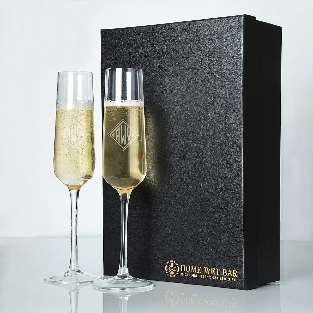 Two champagne flutes with etched monograms filled with bubbly drink beside a black Home Wet Bar gift box.