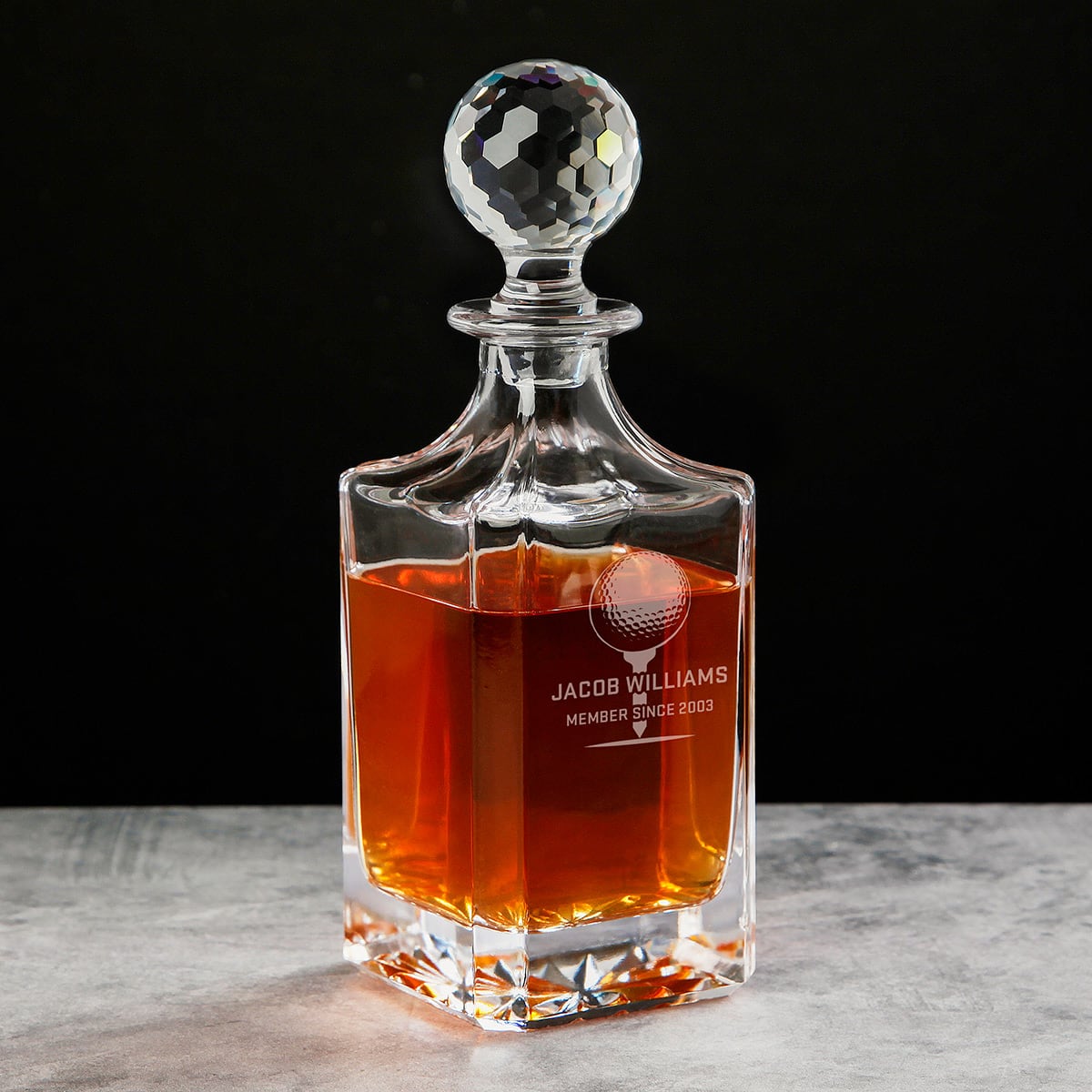 Glass decanter with faceted stopper, filled with amber liquid, etched “JACOB WILLIAMS MEMBER SINCE 2003”.