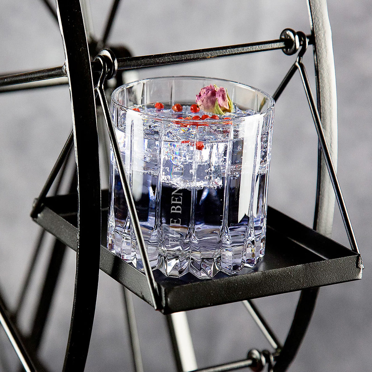 Glass tumbler with clear drink, ice, red berries, and a small flower on a black metal swing tray.