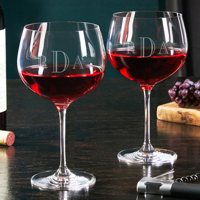 Two stemmed wine glasses filled with red wine on a dark table, with grapes, cork, and corkscrew nearby.