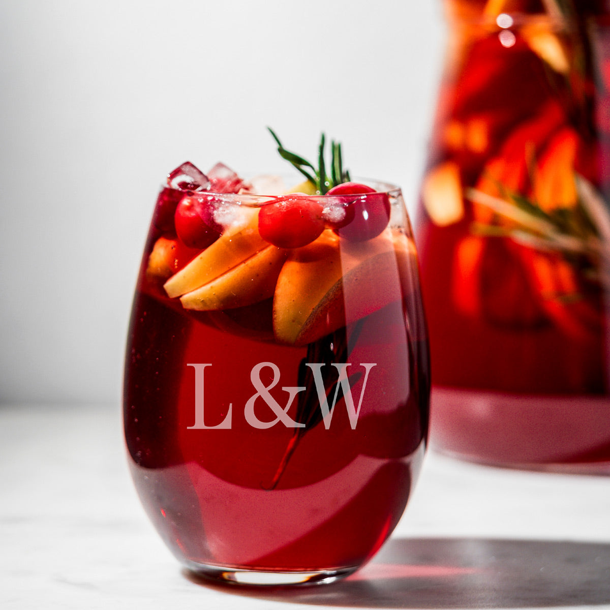Glass of red drink with ice, fruit slices, and cherries; blurred pitcher in background, “L&W” text overlay.