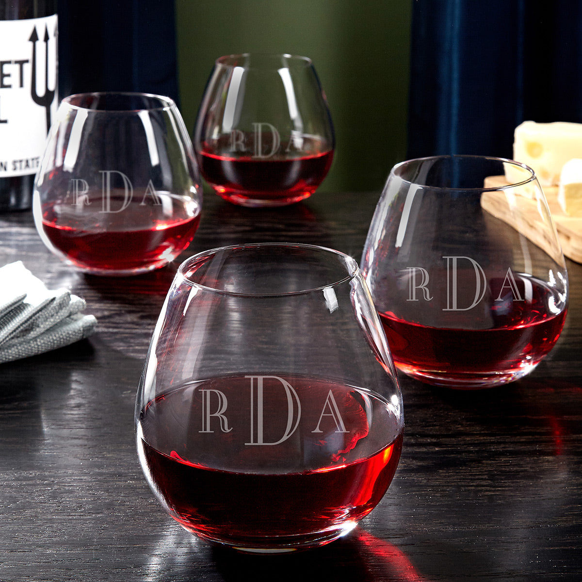 Four stemless wine glasses with red wine and etched “RDA” initials on a dark tabletop.