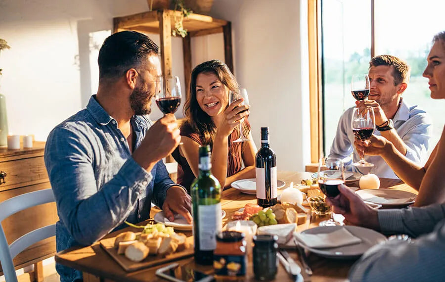Four people at a dining table raising glasses of red wine, with bottles and appetizers in sunlight.