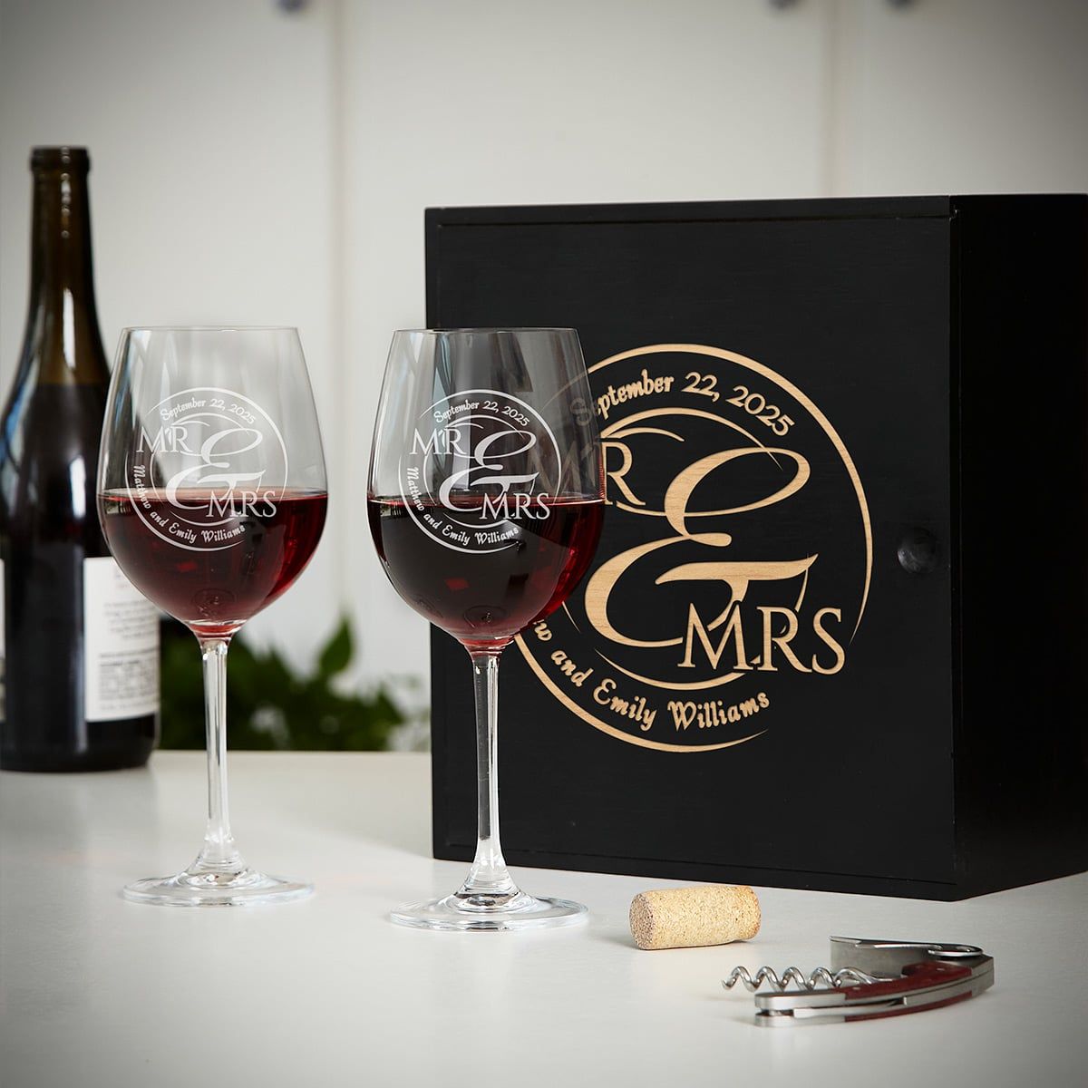 Two engraved wine glasses with red wine in front of a black “Mr & Mrs” gift box, cork and corkscrew on table.
