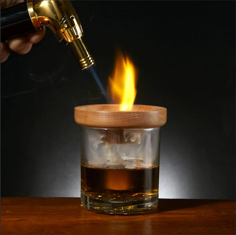Blowtorch flame heats a wooden lid on a glass of dark liquid, creating a burst of fire on top.