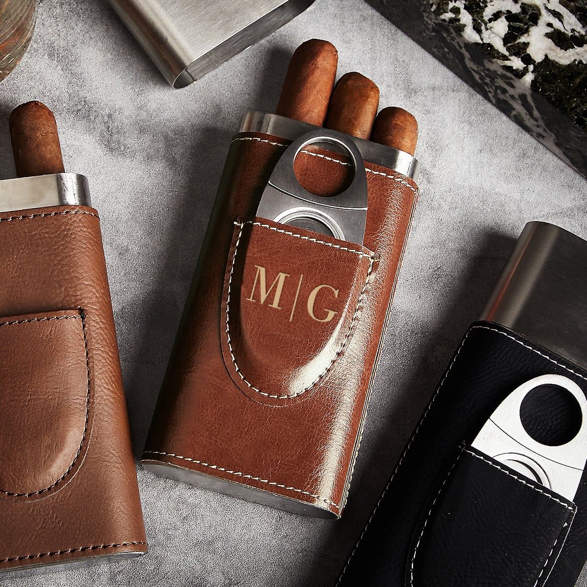 Brown leather cigar case with three cigars and metal cutter, monogrammed “M/G”, on a gray surface.