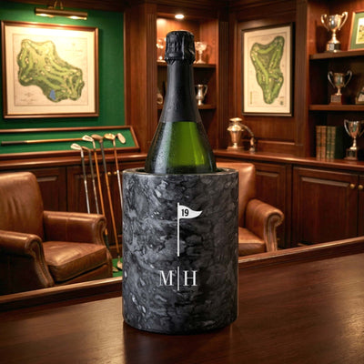 A bottle chilling in a black marble wine chiller with a golf flag icon and monogram, set on a wooden table in a golf-themed lounge with leather chairs and framed course maps