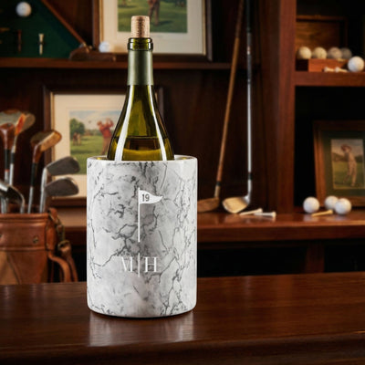 A bottle in a white marble wine chiller engraved with a golf flag icon and initials, set on a wooden table with golf clubs, golf balls, and framed golf artwork in the background