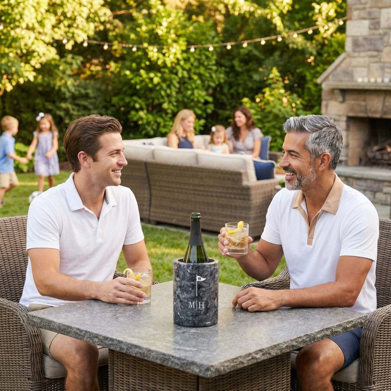 Two men sitting at a stone patio table in a backyard, smiling and holding cocktails, with a bottle in a marble black wine chiller on the table