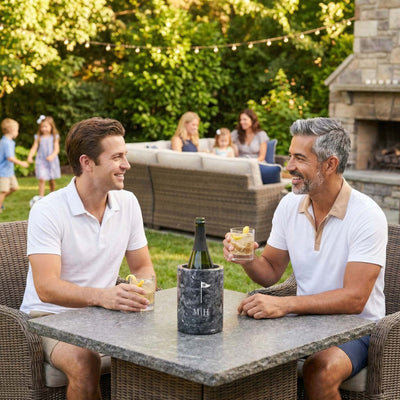 Two men sitting at a stone patio table in a backyard, smiling and holding cocktails, with a bottle in a marble black wine chiller on the table