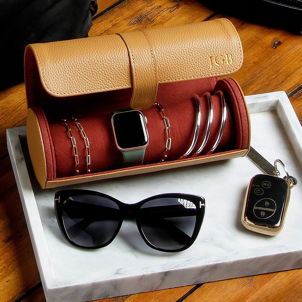Tan leather jewelry roll with bracelets and smartwatch, on a marble tray beside sunglasses and a car key fob.