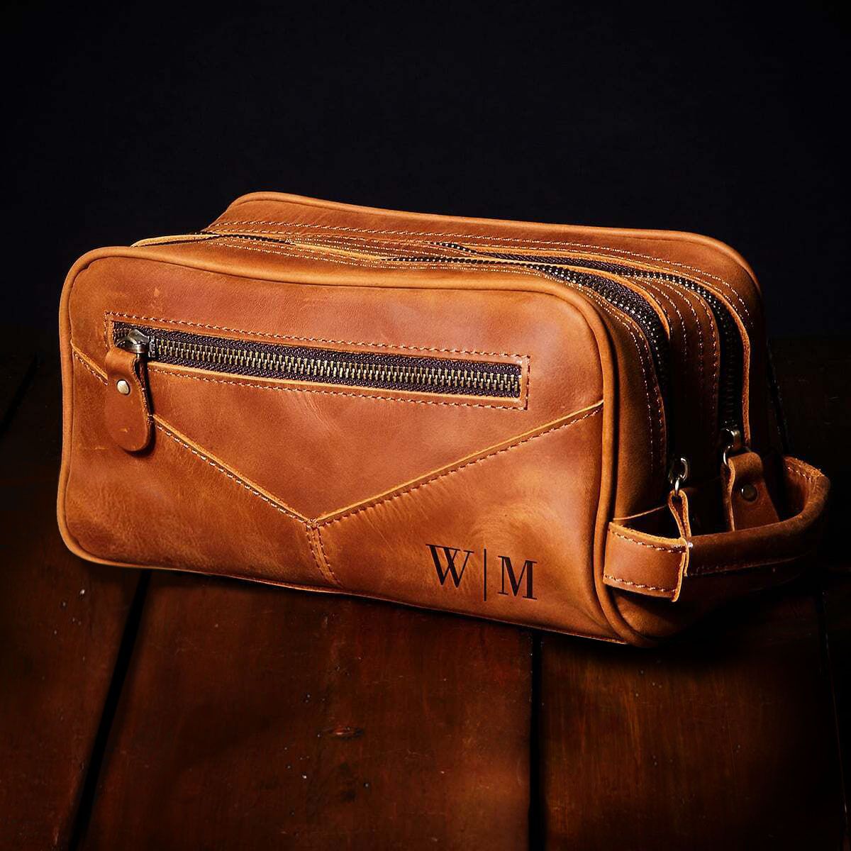 Brown leather toiletry bag with front zipper pocket, multiple compartments, and side handle, on a wooden surface.