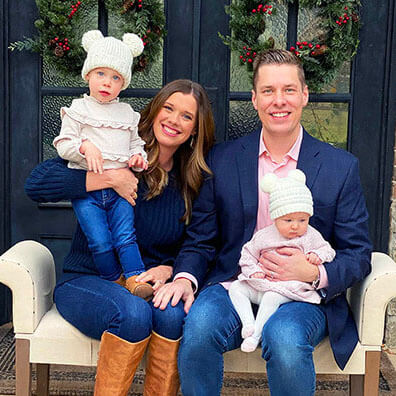 Family of four seated on a bench; parents in center, toddler on mother’s lap and baby on father’s lap, wreaths behind.