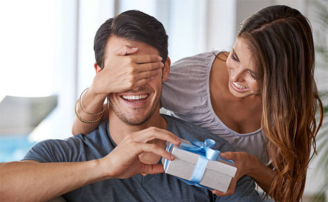 Woman covers a smiling man's eyes while handing him a wrapped gift box with a blue ribbon.