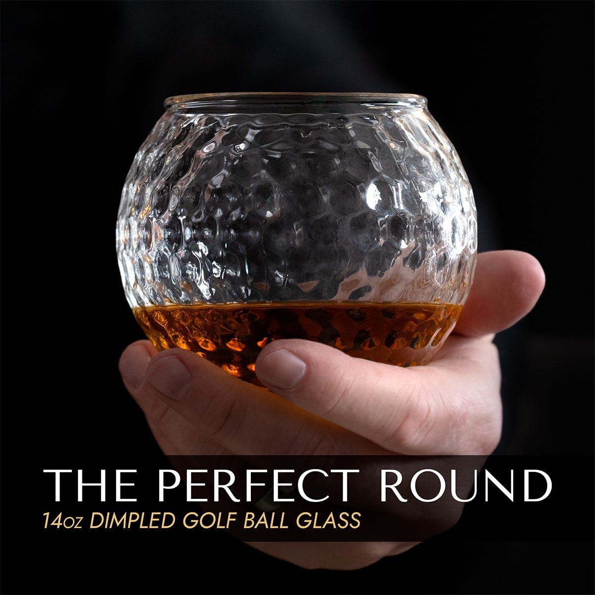 Hand holding a 14 oz round, golf ball inspired glass with whiskey inside