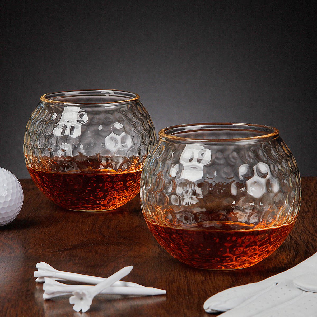 Two clear & round, textured golf ball inspired whiskey glasses filled with liquor on a wooden surface