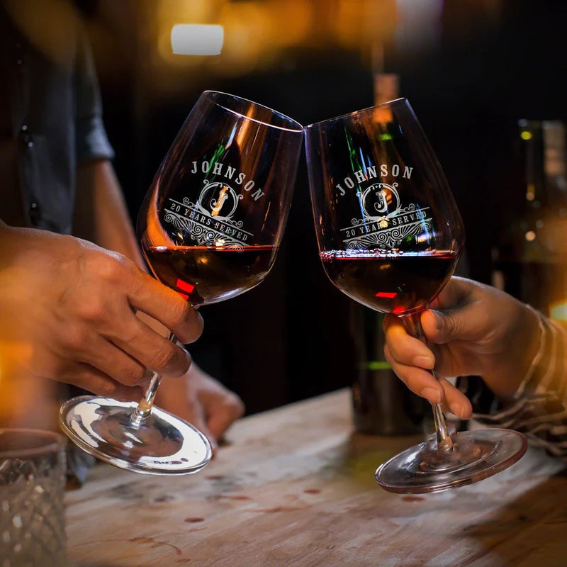 Two hands clinking wine glasses with red wine, “Johnson” logo on glasses, on a wooden table in dim light.