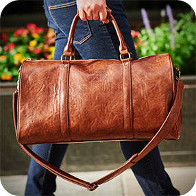 Person carrying a brown leather duffel bag with shoulder strap while walking outdoors