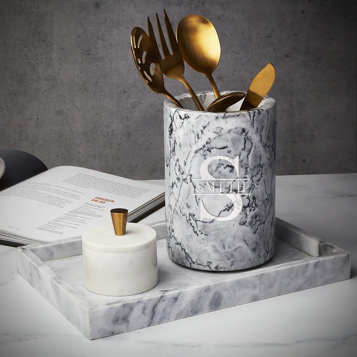 Marble utensil holder with gold utensils and a small lidded jar on a marble tray beside an open menu.